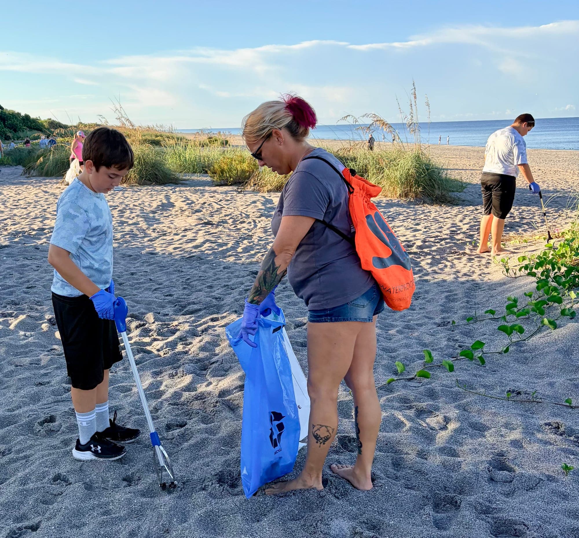 A woman and a boy with litter grabbers fill blue bags on a sandy beach path through the dunes, another volunteer working behind them by the water.
