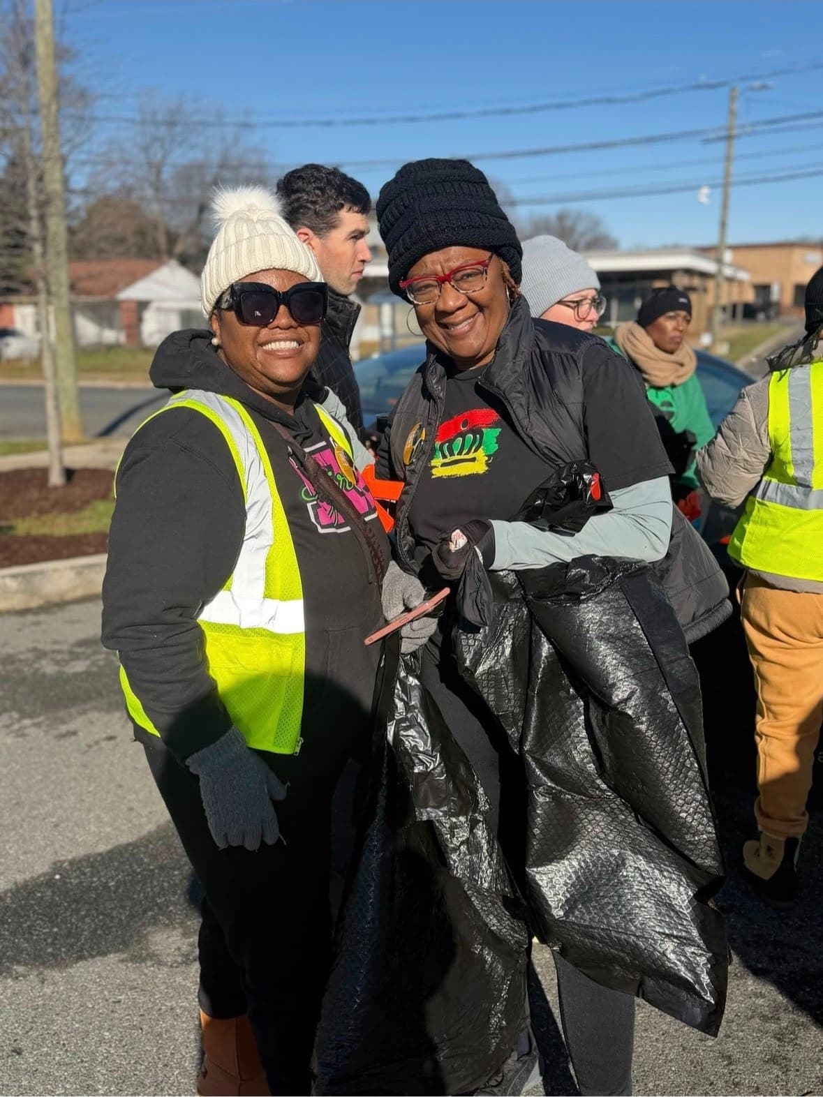 Two smiling volunteers in winter hats hold a black trash bag at a street cleanup, other volunteers gathered behind them.