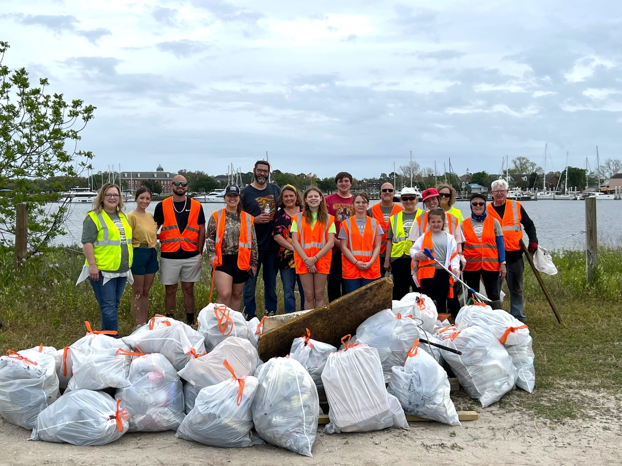 About twenty volunteers in orange and yellow safety vests stand behind a large pile of full white trash bags on a waterfront, a marina of sailboats behind them.