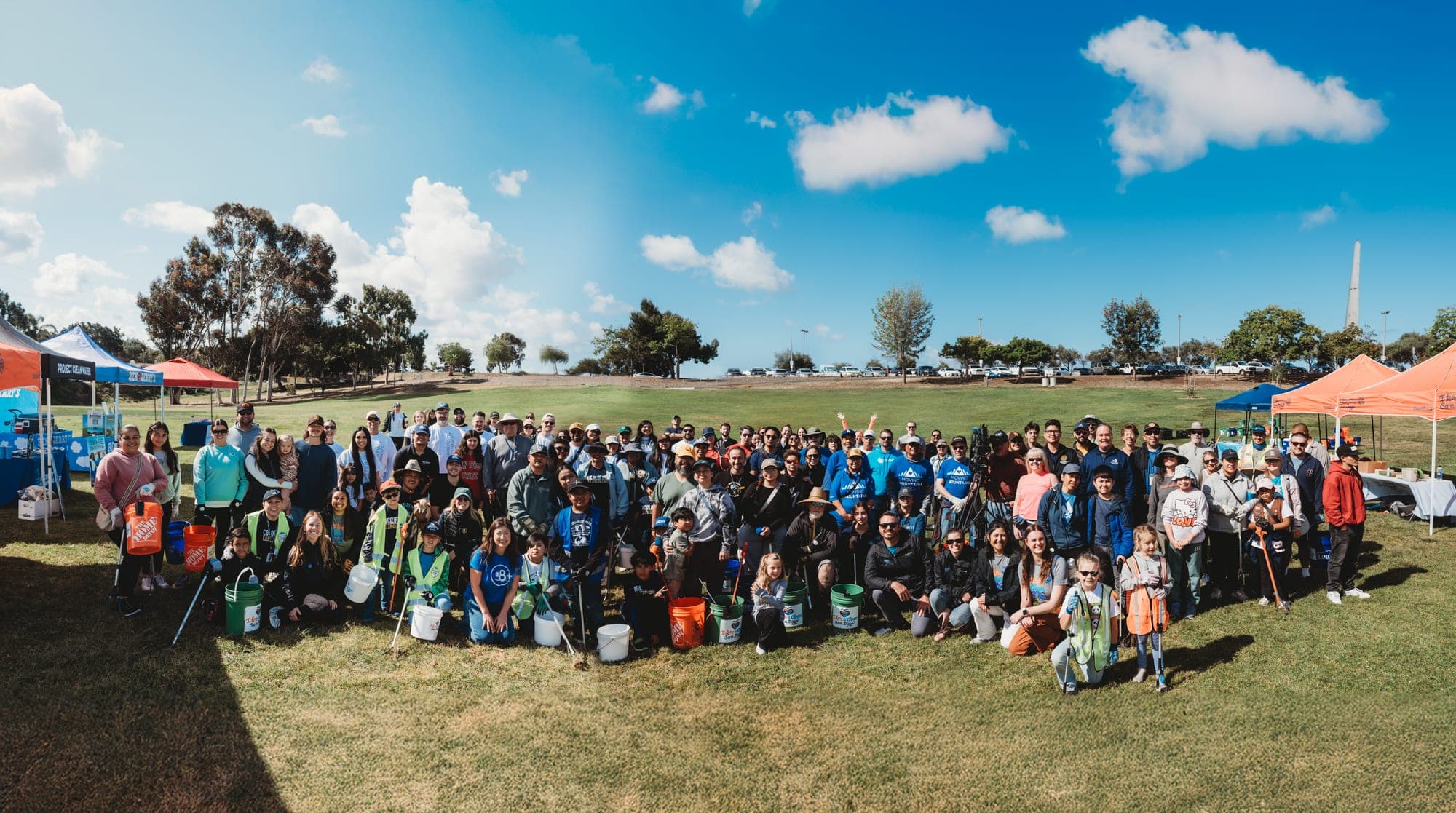 More than a hundred volunteers pose together on a wide park lawn after a cleanup, buckets and litter grabbers at their feet, event tents on either side under a bright blue sky.