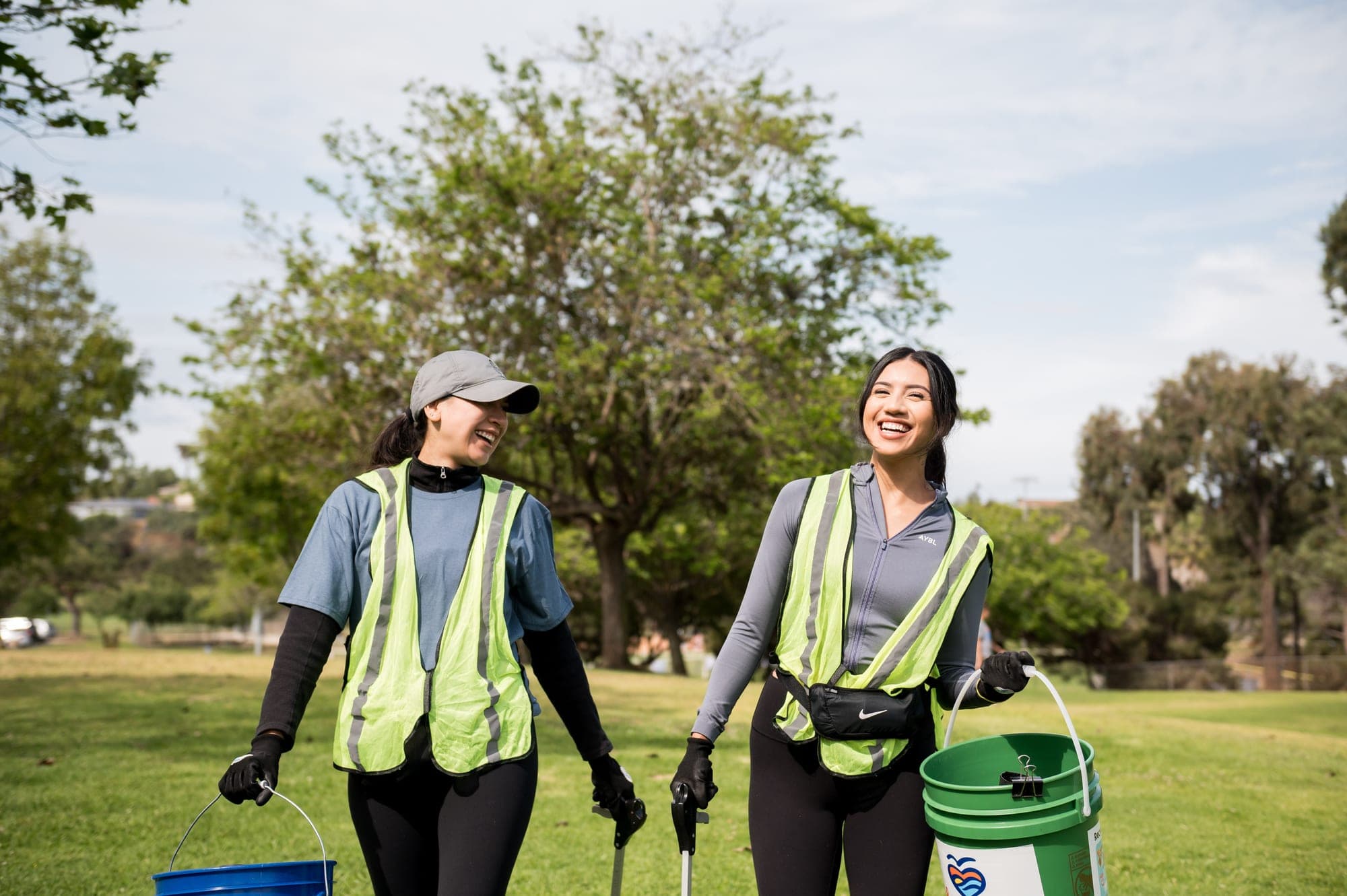 Two volunteers in yellow safety vests laugh as they walk across a park lawn carrying buckets and litter grabbers, trees behind them.