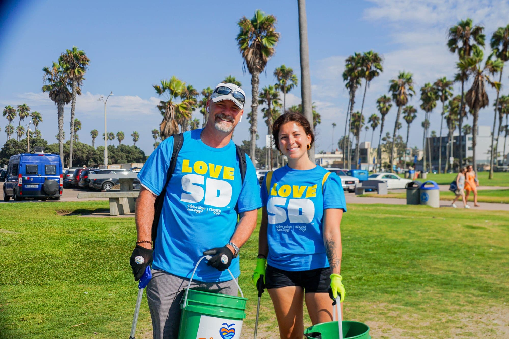 Two volunteers in blue Love SD t-shirts smile on a park lawn lined with palm trees, holding green buckets and litter grabbers.