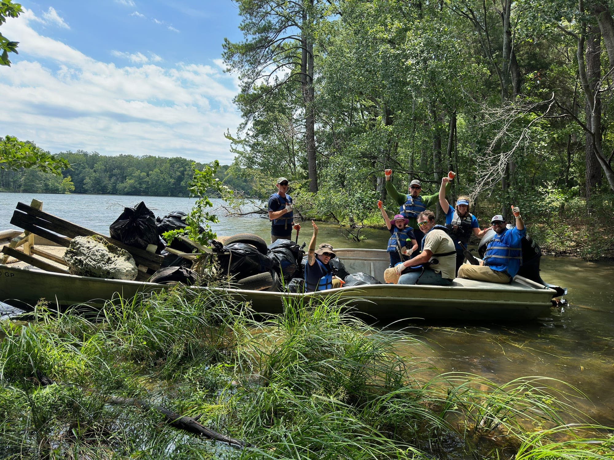 Volunteers in life jackets cheer from a flat-bottomed boat piled with trash bags, timber and tires pulled from a wooded lake shore.