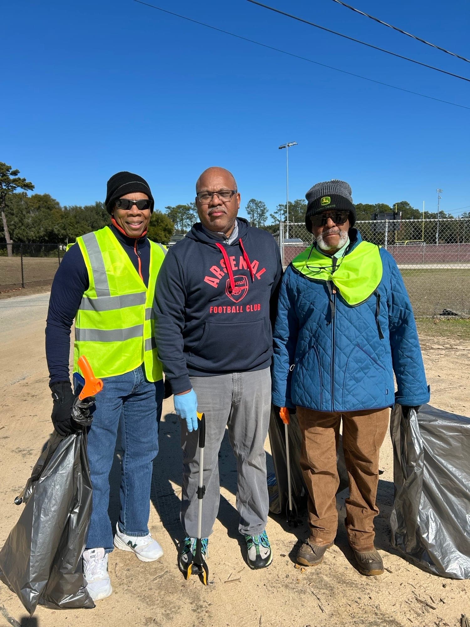 Three volunteers in winter hats and safety vests stand side by side on a sunny path, each holding a full trash bag and a litter grabber.