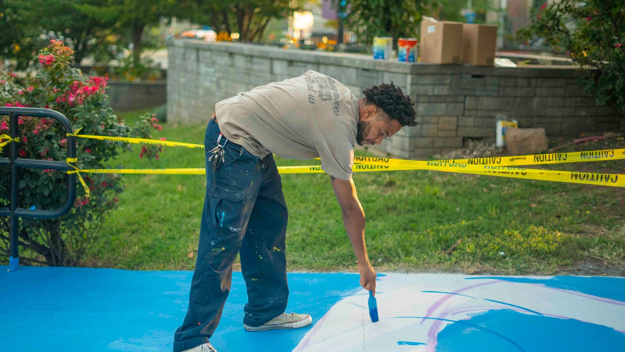 A volunteer in a paint-spattered t-shirt bends to brush blue paint onto a ground mural taped off with caution tape, a rose bed and a stone wall behind him.