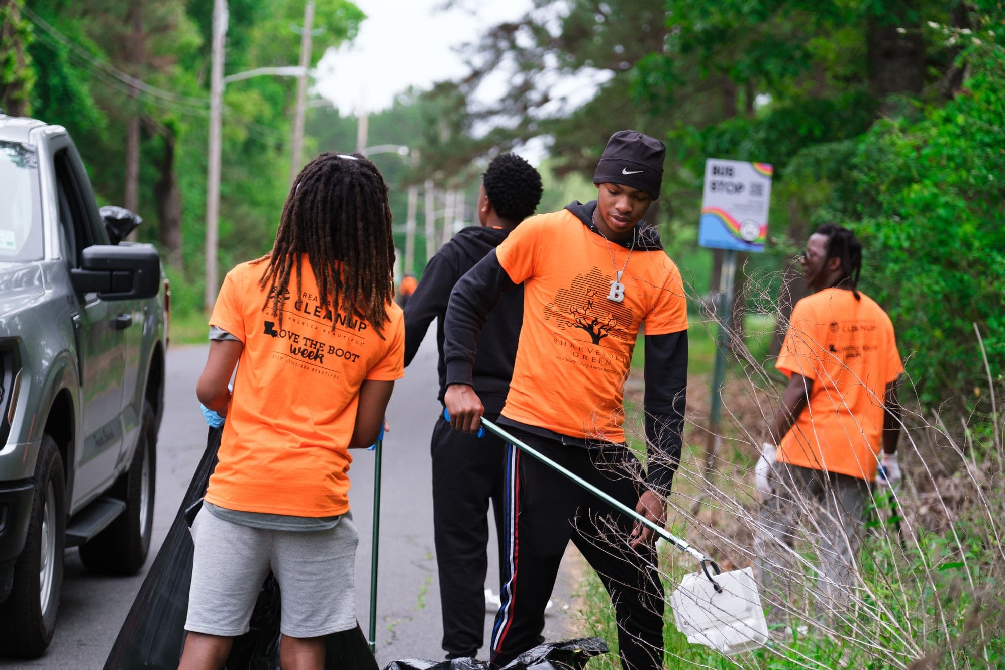 Young volunteers in orange Great American Cleanup t-shirts work a roadside verge with litter grabbers and black trash bags, a pickup truck parked beside them.
