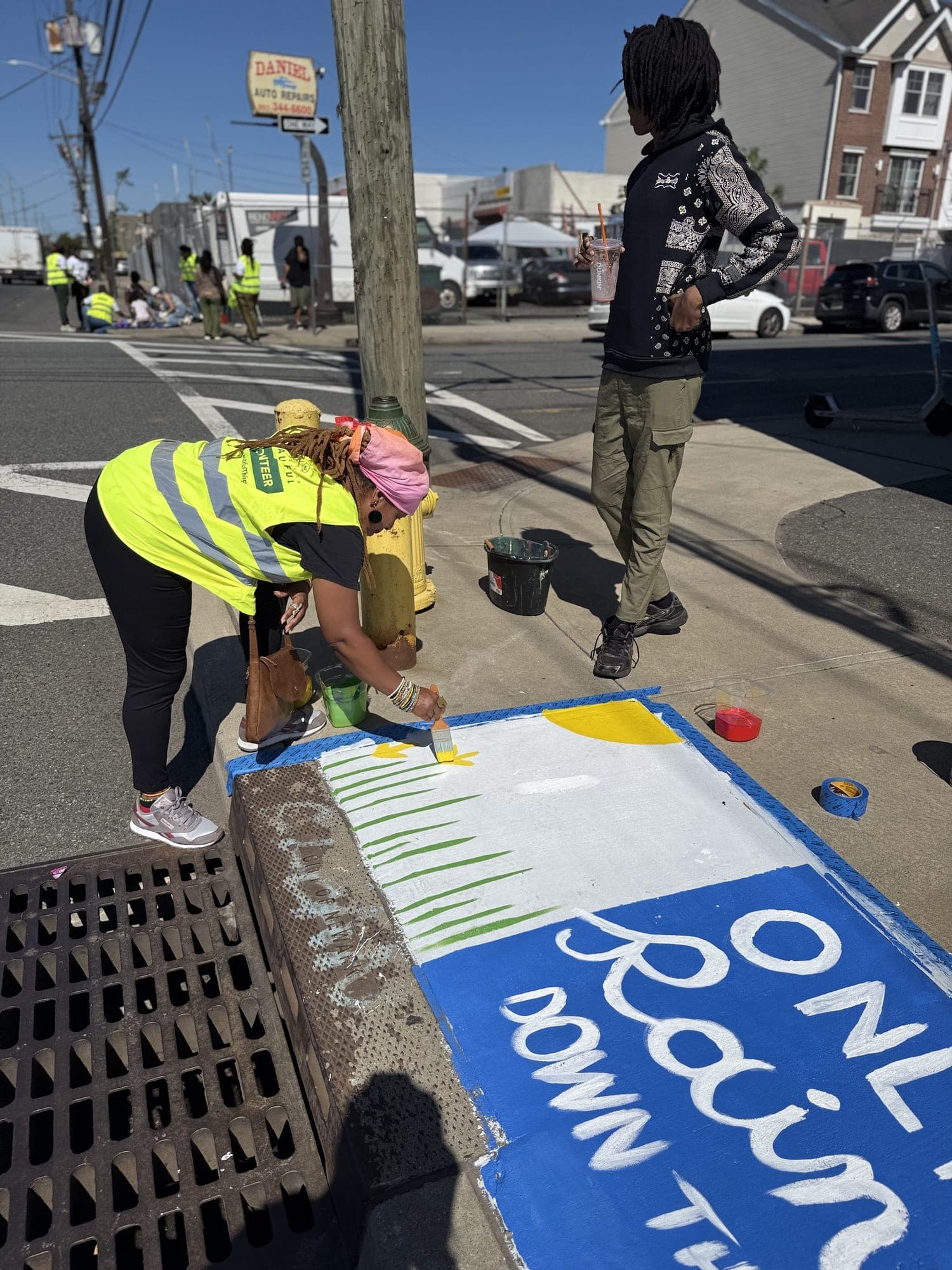 A volunteer in a yellow vest paints a storm-drain mural reading “Only rain down the drain” on a city sidewalk while another looks on.