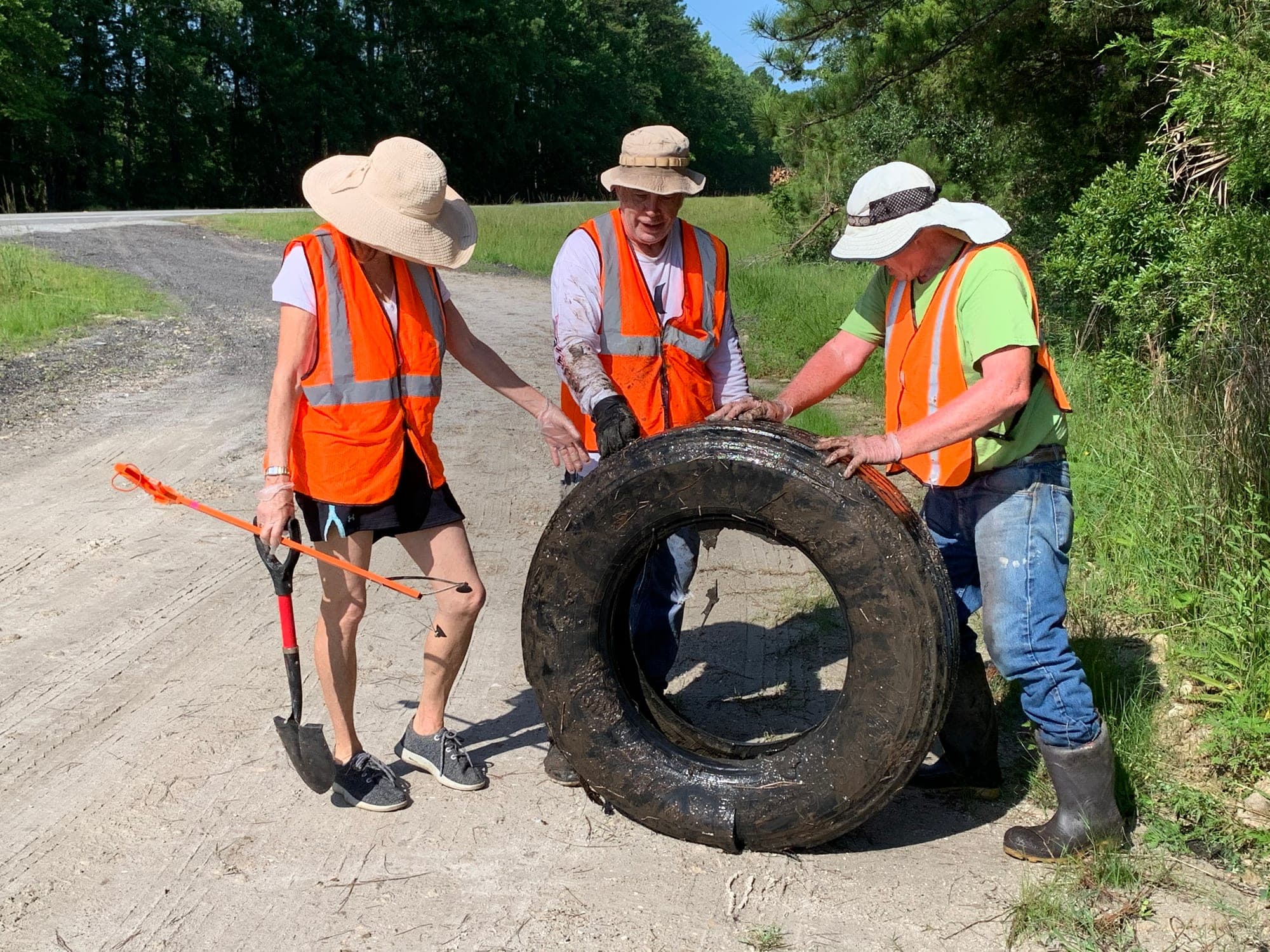 Three volunteers in orange safety vests and sun hats steady a large muddy tire on a gravel road, one holding a shovel and a litter grabber.