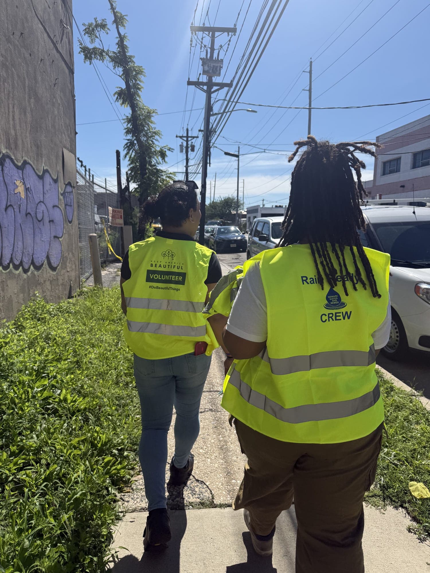 Two volunteers in yellow safety vests, one reading Keep America Beautiful Volunteer, walk away down a sunny city sidewalk lined with shrubs and graffiti.