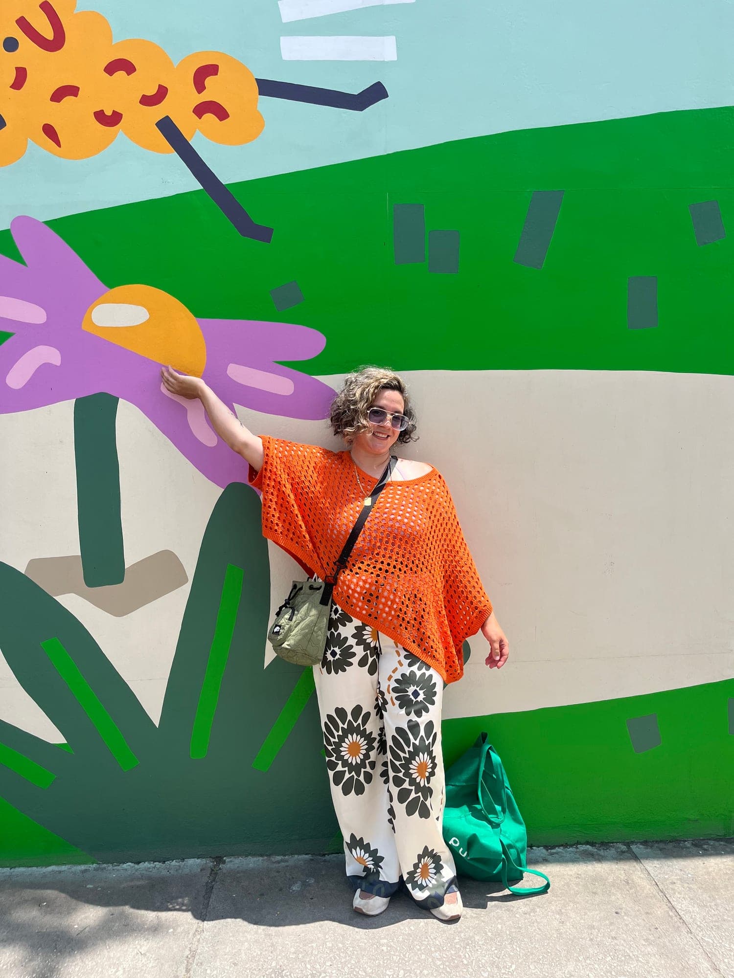 A volunteer in an orange top leans against a bright mural of giant flowers on a city wall, one arm raised to a painted bloom.