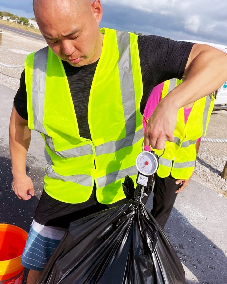 A volunteer in a yellow safety vest weighs a full black trash bag on a hand-held scale at a cleanup.