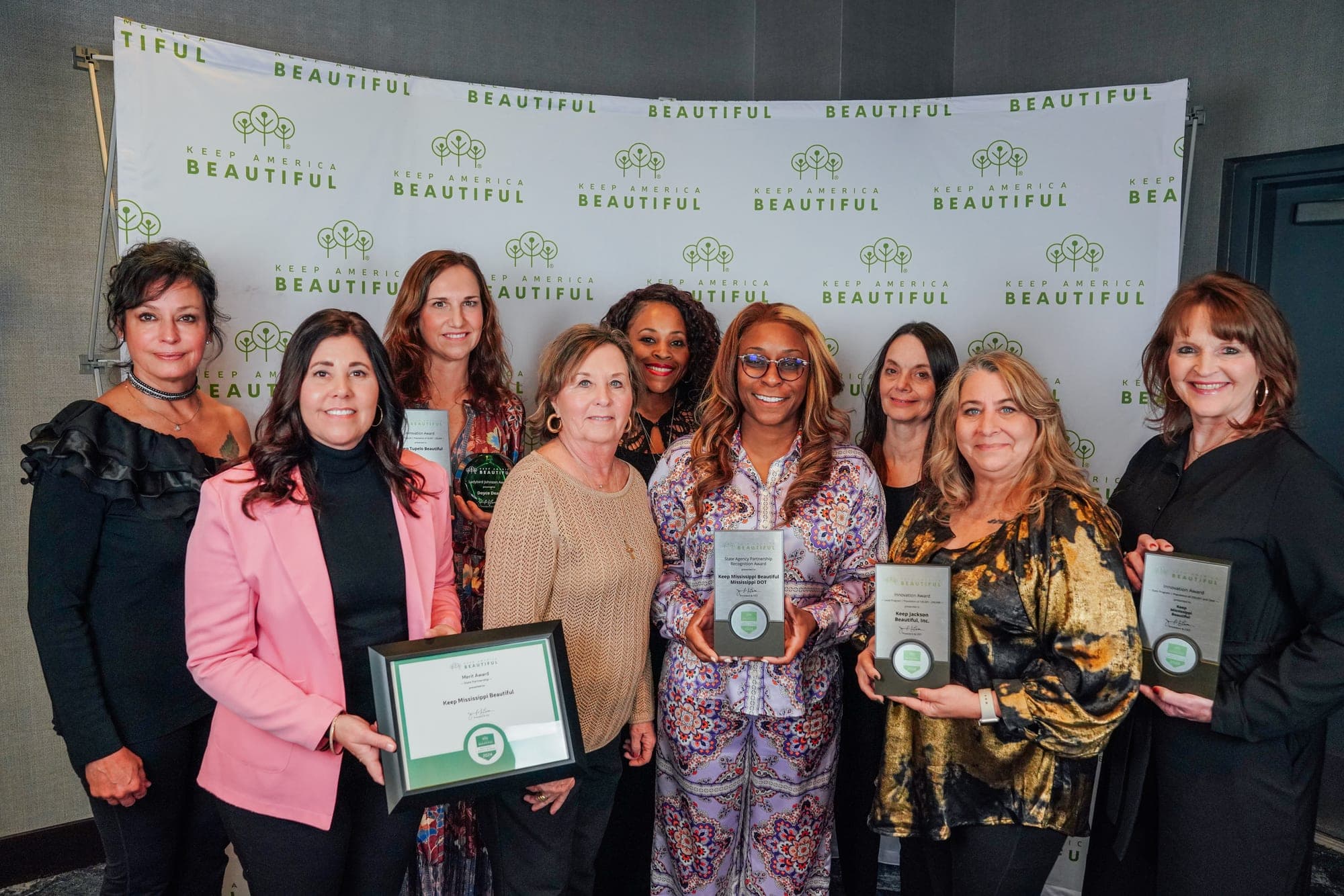 A group of ten women pose together before the Keep America Beautiful backdrop, several holding award plaques and one a framed certificate.