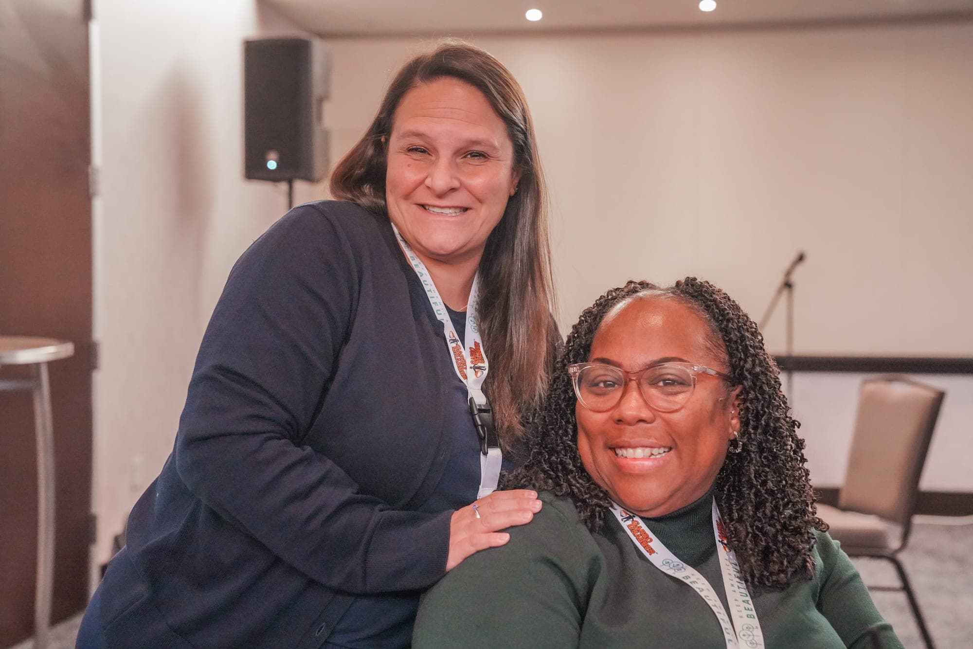 Two smiling conference attendees wearing lanyards pose together, one resting a hand on the other's shoulder.