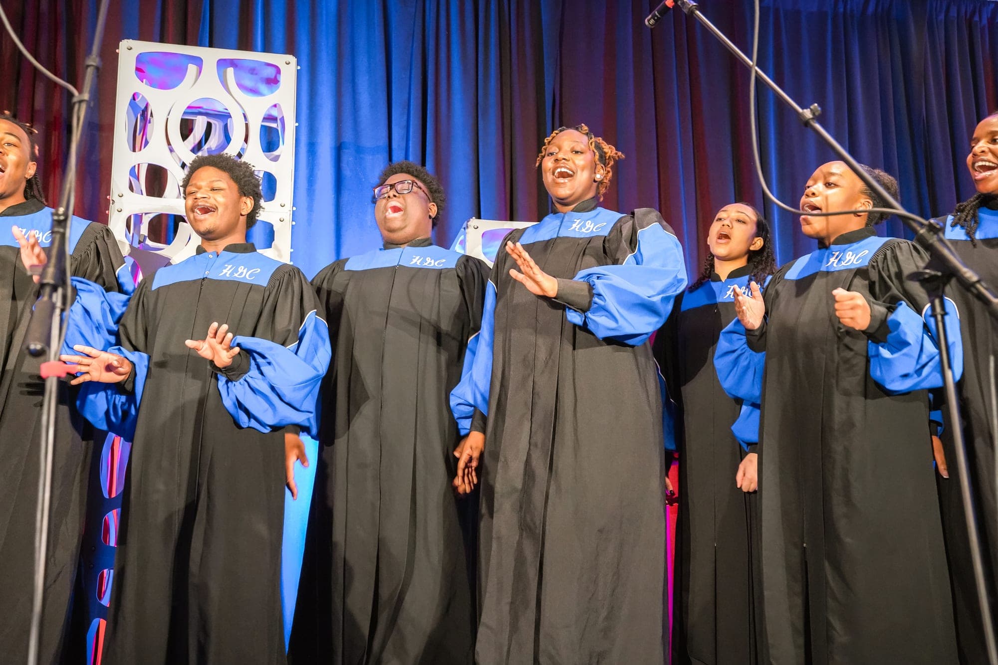 A gospel choir in black and blue robes sings on stage, several members with raised hands and open mouths mid-song.