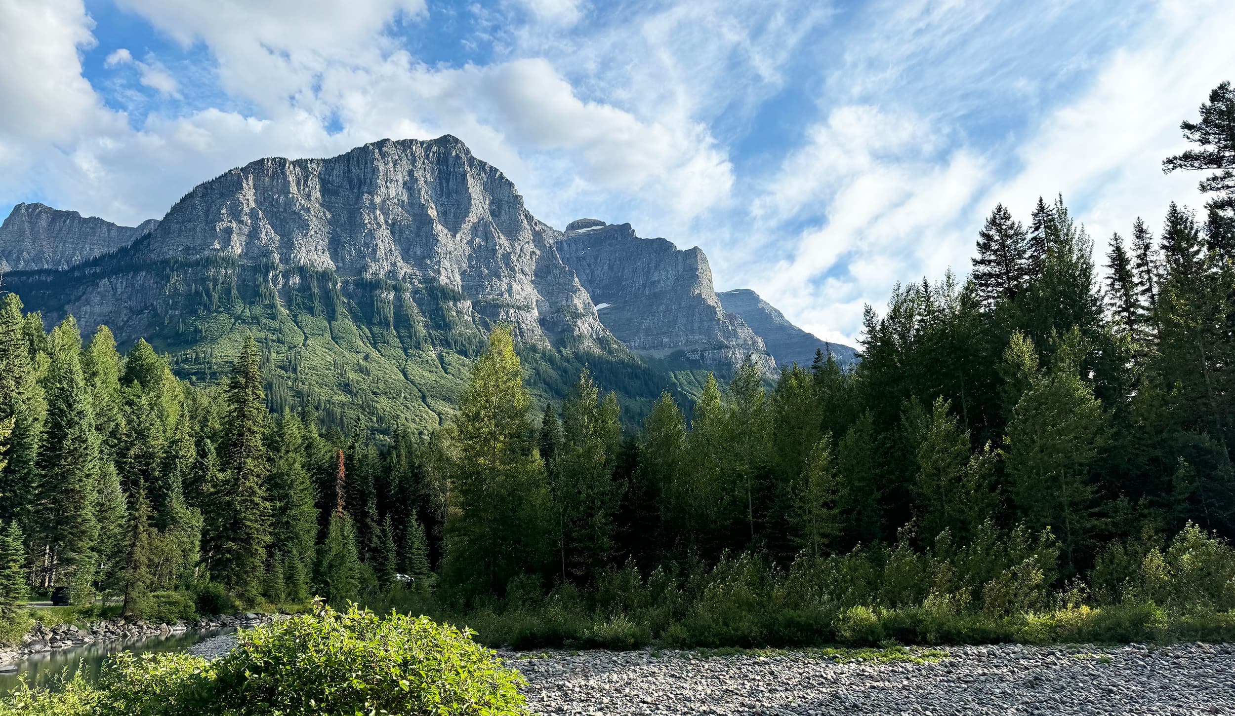 A grey limestone peak in Grand Teton National Park rises over a dense conifer forest, with a braided gravel riverbed in the foreground under a blue sky of broken cloud.
