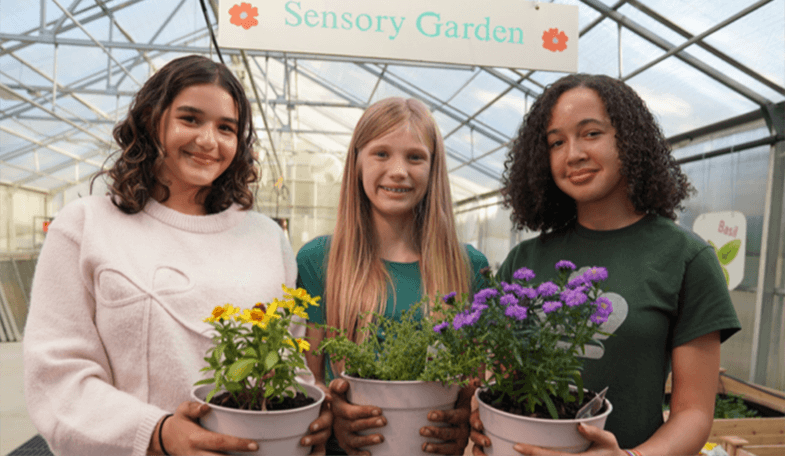 Three students stand side by side in a greenhouse beneath a “Sensory Garden” sign, each holding a potted plant in flower — marigolds, herbs and purple asters.