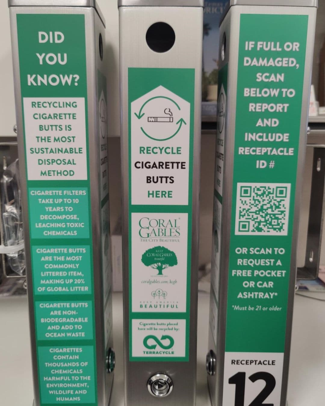 Three Keep Coral Gables Beautiful cigarette receptacles standing side by side, their panels printed with recycling facts, a QR code for reporting a full or damaged unit, and a receptacle ID number.