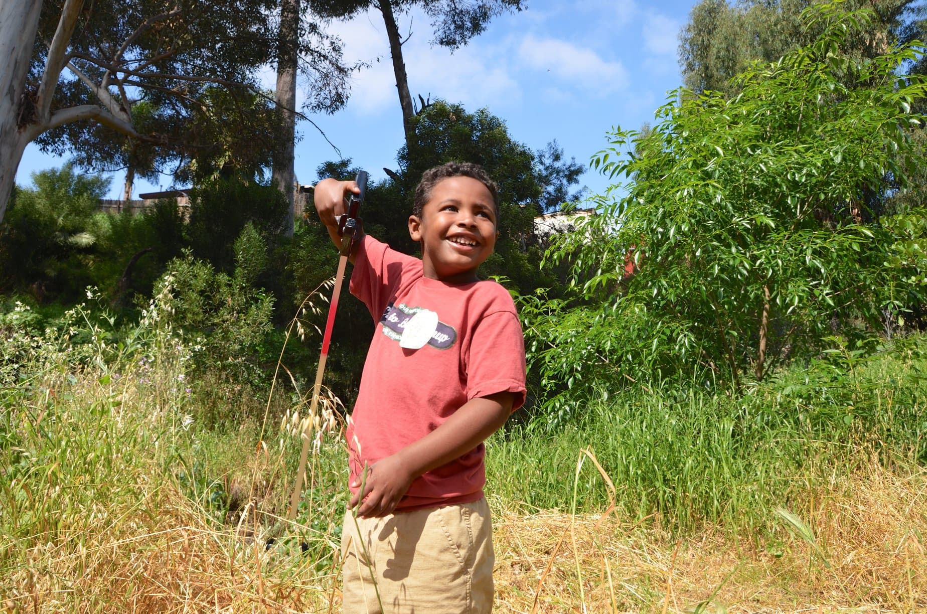 A smiling boy in a red t-shirt stands in tall grass at a Creek to Bay cleanup, holding a litter grabber up high, trees and green brush behind him.
