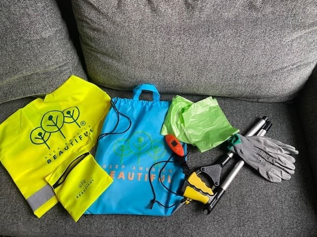 A Great American Cleanup volunteer kit laid out on a gray sofa: a lime hi-vis vest with a silver reflective stripe and the Keep America Beautiful tree logo, a blue Keep America Beautiful drawstring bag, a black and yellow long-handled litter grabber, a bundle of bright green liner bags, gray work gloves, and a small orange digital hanging scale.