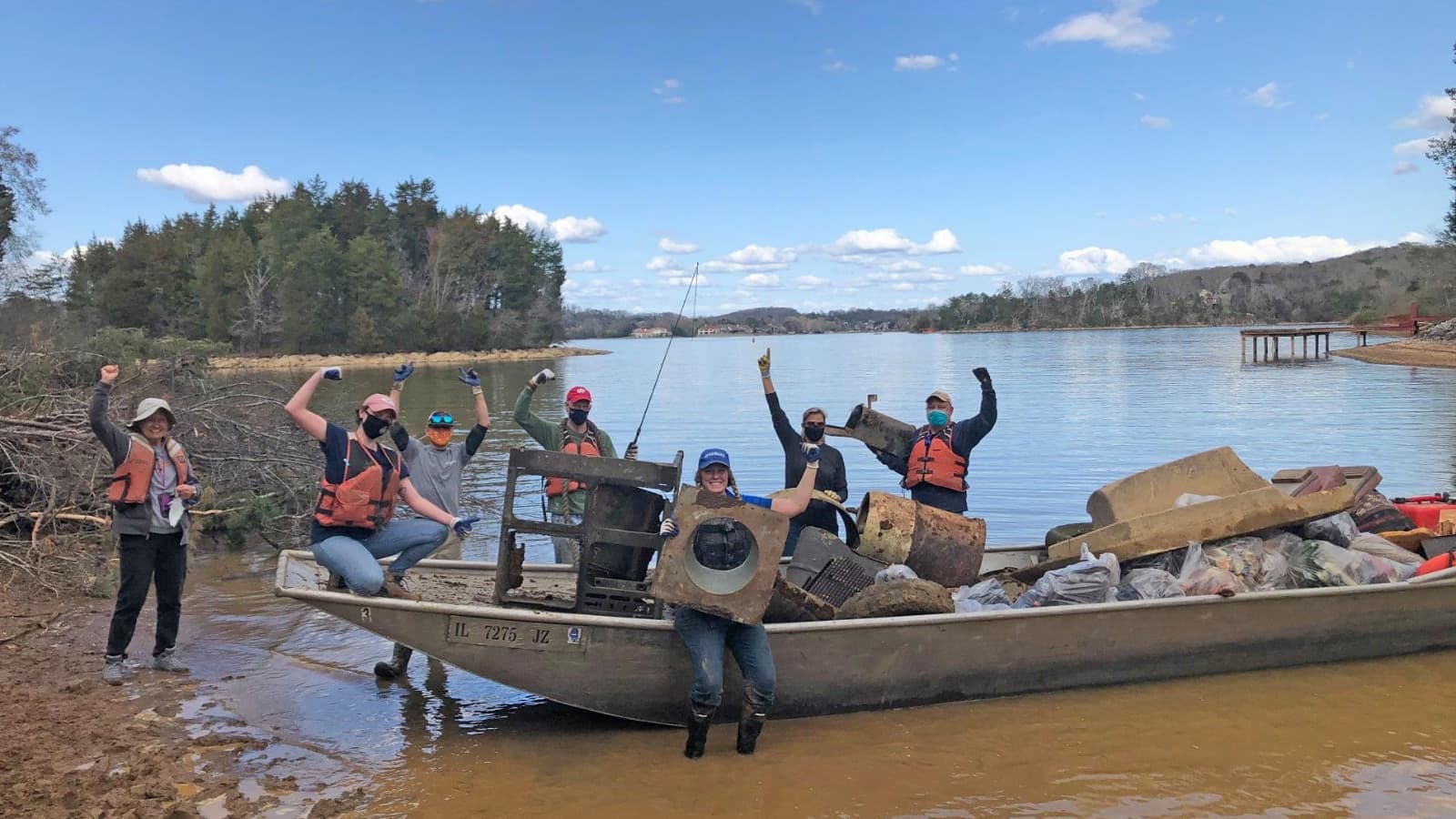 Volunteers in life vests and work gloves cheer with raised arms beside a flat-bottomed boat heaped with bagged trash, a rusted metal frame, barrels and scrap pulled from a lake, a wooded shoreline behind them.
