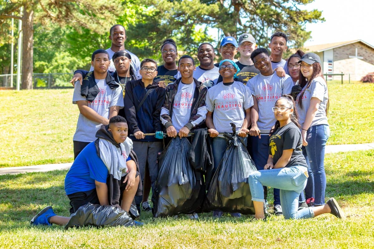 About fifteen high-school students in gray Keep Conyers-Rockdale Beautiful t-shirts stand and kneel together on a sunlit school lawn behind two full black trash bags.