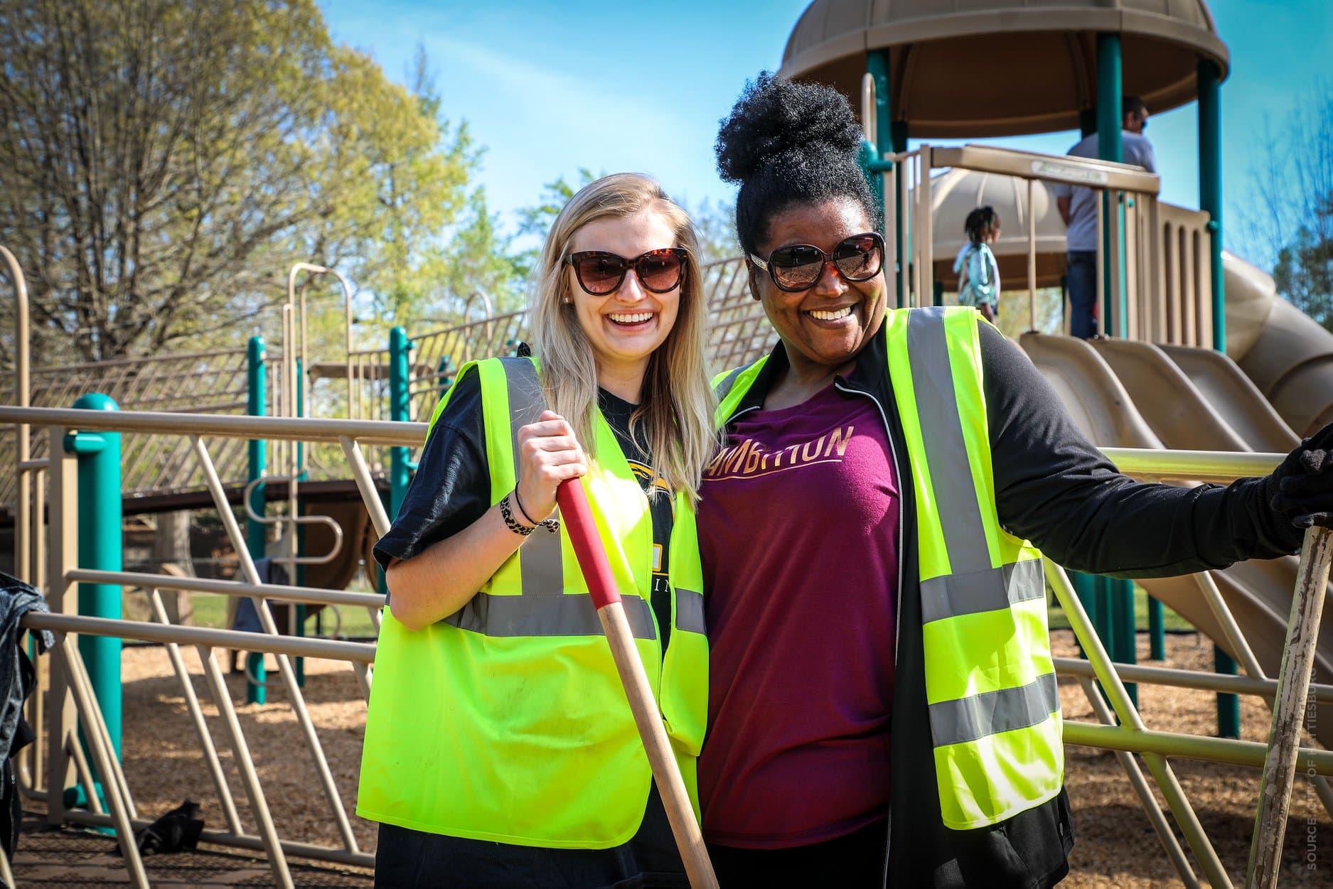 Two Keep Hattiesburg Beautiful volunteers in hi-vis vests and sunglasses stand arm in arm at a playground, leaning on rakes, with children playing on the climbing frame behind them.