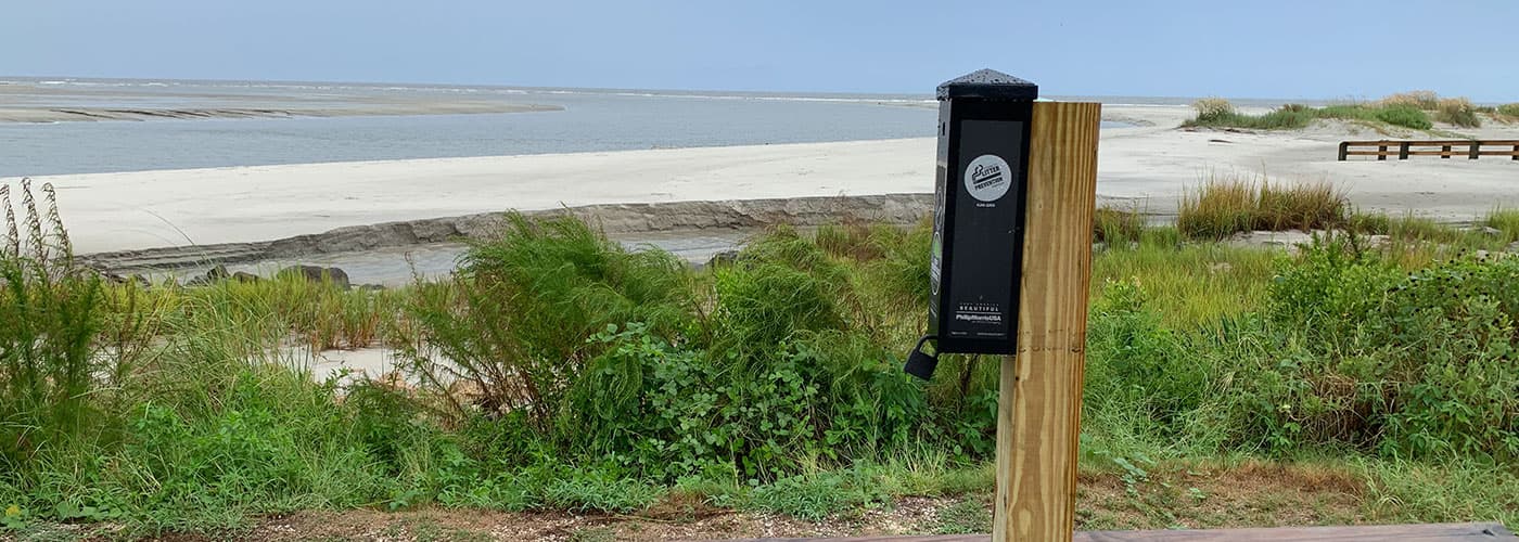 A black cigarette-litter receptacle mounted on a wooden post at the edge of a beach, dune grasses around it and open water behind.