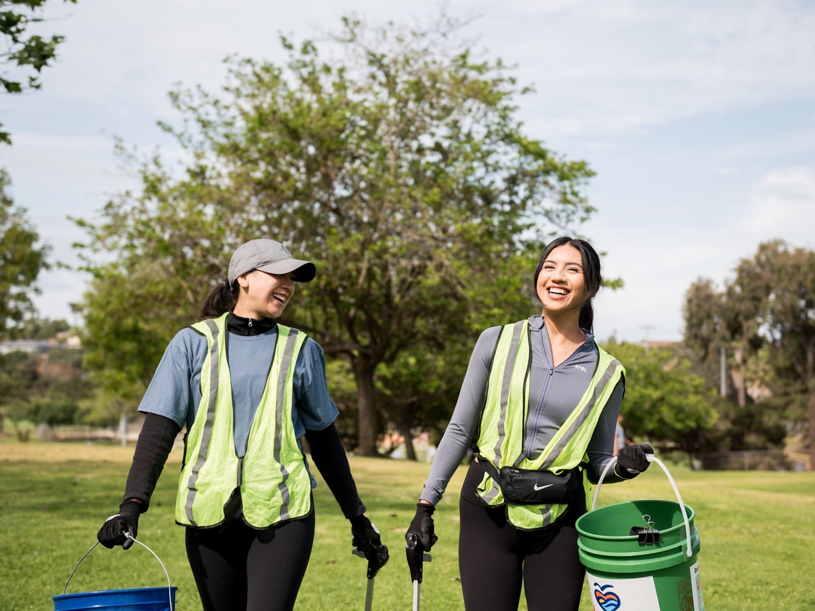 Two volunteers in yellow safety vests and work gloves laugh as they walk across a sunny park lawn carrying buckets and litter grabbers.