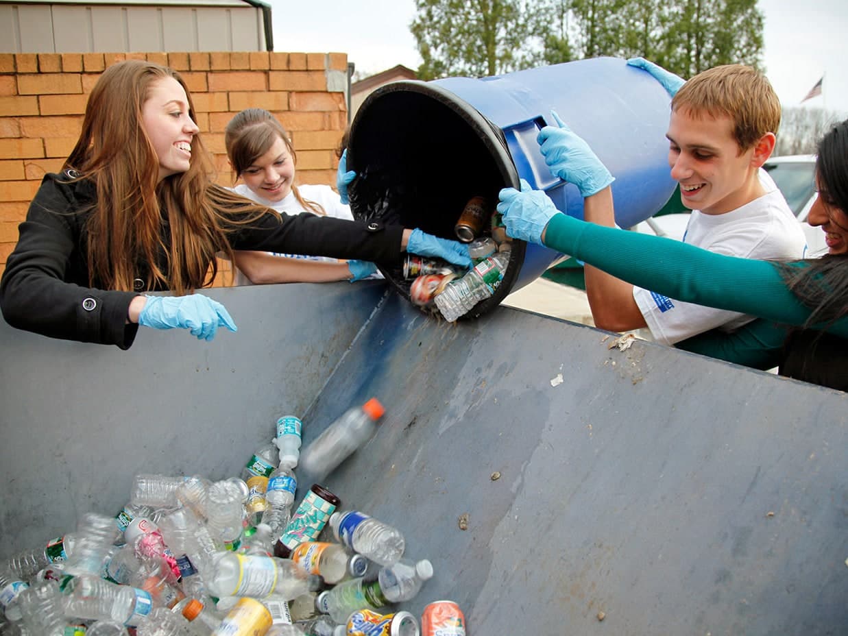 Four smiling young volunteers in blue gloves tip a blue bin of bottles and cans into a large recycling container.